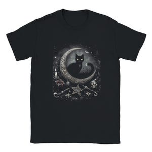 May include: A black short-sleeved t-shirt featuring a graphic print of a black cat sitting on a large, ornate crescent moon. The design includes a lit candle, a small gift box, a star-shaped ornament, and various celestial elements in shades of grey.