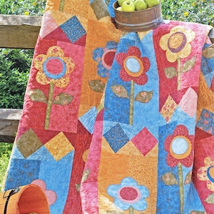 Può includere: Un quilt colorato con un design a patchwork che presenta grandi fiori stilizzati in tonalità di rosso, blu, giallo e verde. Il quilt è drappeggiato su una recinzione di legno e un cesto di mele verdi si trova in primo piano.