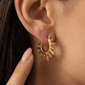 Puede incluir: Pendientes de aro dorados con un diseño de rayos de sol. Los pendientes tienen forma de semicírculo con rayos puntiagudos que se extienden hacia afuera. Las joyas se llevan en la oreja, destacando los intrincados detalles del diseño solar.