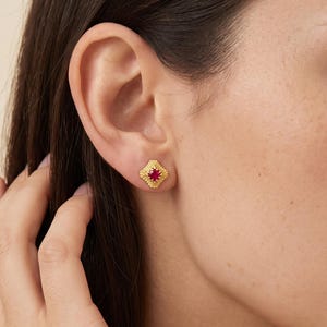 Könnte beinhalten: Ein kleiner goldener Ohrstecker mit einem texturierten, rautenförmigen Rahmen und einem leuchtend roten Edelstein in der Mitte. Die Metalloberfläche hat eine feine, körnige Struktur und der Ohrstecker wird mit einem Stift befestigt.
