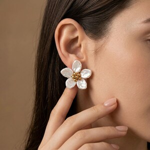 May include: A white, flower-shaped earring with five petals and a cluster of small gold beads at the centre. The earring is worn on the ear, highlighting its delicate design and texture. The background is a neutral, warm tone.