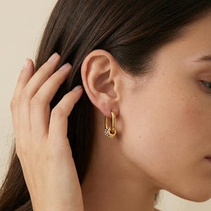 Puede incluir: Pendientes de aro dorados con forma rectangular y un pequeño dije redondo adornado con diminutos cristales. Los pendientes se lucen en la oreja de una persona, mostrando su diseño y brillo.