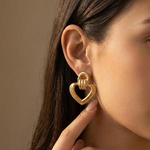 May include: Gold-colored heart-shaped earrings. The earrings have a layered design with a heart outline and a small loop at the top. The earrings are a shiny gold color and are worn on the ear.
