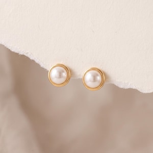 May include: A pair of stud earrings featuring a round, white pearl set in a smooth, polished gold-toned metal frame. The earrings have a circular design with a raised center, showcasing a classic combination of lustrous white pearl and gold.