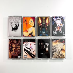 Peut inclure: Huit cassettes audio du groupe Korn disposées sur deux rangées. Les pochettes présentent des illustrations graphiques distinctes, notamment une poupée de chiffon, un enfant sur une balançoire, une foule de visages et des images surréalistes.