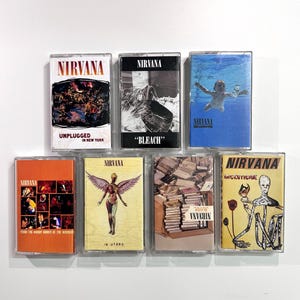 Peut inclure: Une collection de sept cassettes Nirvana. Les cassettes présentent des pochettes d'album avec divers designs, dont un bébé dans l'eau, une figure ailée et des textes tels que "Unplugged in New York" et "In Utero".