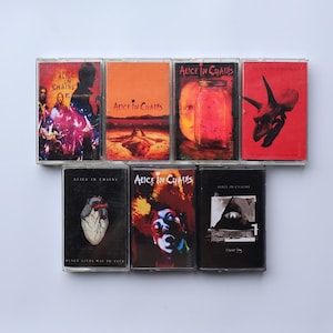 Könnte beinhalten: Sieben Musikkassetten mit Albumcovern der Band Alice in Chains. Die Cover zeigen verschiedene Designs, darunter Porträts und abstrakte Kunst. Das Logo der Band ist auf einigen Kassetten zu sehen. Auf weißem Hintergrund.