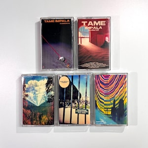 May include: Five Tame Impala cassette tapes are displayed. The album art varies, with designs such as a black background with a red line, a desert scene, and a psychedelic pattern. The text "Tame Impala" is printed on each cassette, showcasing the band's branding.