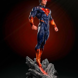 May include: A superhero statue in a dynamic pose, clad in a blue and red suit. The figure stands atop a rocky base, with glowing red accents on the chest, arms, and legs. The background is a gradient of red and black.