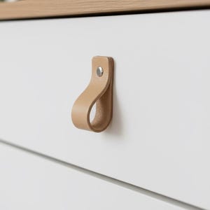 Op de afbeelding: Close-up van een witte dressoir met twee lades, voorzien van bruin lederen lusgrepen. De grepen zijn bevestigd met zilveren klinknagels. De bovenkant van de dressoir is licht hout.