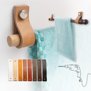 Könnte beinhalten: Zwei Leder-Handtuchhalter mit Holzdübeln, einer mit einem hellbraunen Lederriemen und der andere mit einem dunkelbraunen Holzdübel. Eine Auswahl an Lederfarbmustern wird unten angezeigt. Zwei türkisfarbene Handtücher hängen an den Haltern.