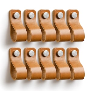 Può includere: Dieci maniglie per mobili in pelle marrone con rivetti in metallo color argento sono disposte in due file da cinque. Le maniglie hanno un design ad anello per una facile presa, su sfondo bianco.