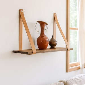 Peut inclure: Une étagère flottante en bois avec des lanières de cuir, supportant deux vases décoratifs en céramique. L'étagère est en bois foncé et les lanières de cuir sont de couleur marron clair. Les vases sont marron et orange.