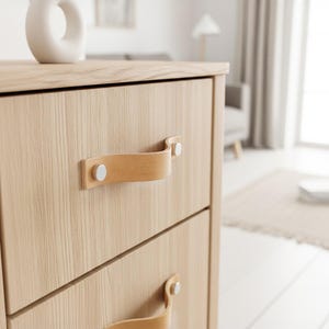 Op de afbeelding: Een licht houten dressoir met twee lades. Elke lade heeft een lichtbruine leren handgreep die met zilverkleurige klinknagels is bevestigd. Bovenop het dressoir staat een witte vaas. De kamer heeft witte muren en een neutraal tapijt.