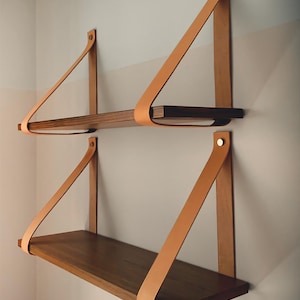 Peut inclure: Deux étagères flottantes en bois avec des planches en bois foncé et des lanières en cuir beige. Les étagères sont fixées sur un mur de couleur claire. Les lanières en cuir sont fixées au mur avec des rivets argentés.