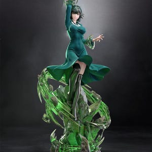 May include: A detailed figurine of a character in a flowing green dress and black boots, with short black hair. The figure is posed dynamically, with one arm raised, surrounded by swirling green energy effects. The base is a rocky structure with glowing green accents.