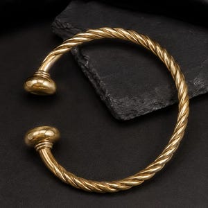 May include: A gold-toned open cuff bracelet featuring a twisted rope-like texture along the band. The bracelet terminates at both ends with smooth, polished, rounded dome-shaped caps that provide a distinct contrast to the twisted metal design.