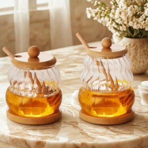 Puede incluir: Dos tarros de miel de vidrio con tapas y cucharas de madera. Los tarros tienen un diseño ondulado único y se asientan sobre bases redondas de madera. La miel en el interior es de color ámbar dorado, y las tapas tienen un pequeño pomo de madera.