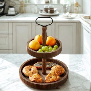Puede incluir: Una bandeja de servicio de madera oscura de dos niveles con un asa negra. El nivel superior contiene naranjas y uvas verdes, mientras que el nivel inferior muestra pasteles y un bagel. La bandeja está diseñada para servir alimentos.