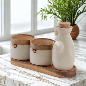 Puede incluir: Un juego de cocina con dos pequeños recipientes beige con tapas de madera y una pequeña botella de aceite beige con tapón de corcho. Los artículos están sobre una bandeja de madera. Una planta verde está en el fondo.