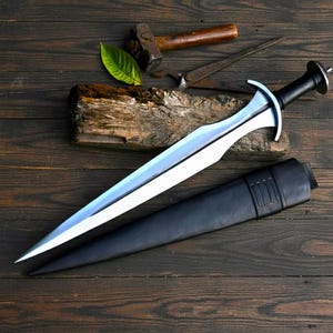 May include: A silver and black sword with a leather sheath on a wooden surface. The sword features a double-edged blade and a crossguard. A hammer, saw, and wood are also visible.