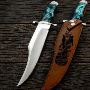 Può includere: Un grande coltello in acciaio lucido con un manico verde acqua e argento, accanto a un fodero in pelle marrone. Il fodero presenta un'immagine incisa di una figura barbuta con un elmo cornuto. Il coltello ha una lama larga e una guardia incrociata argentata.