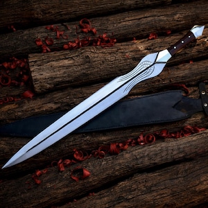 May include: A long, silver sword with a decorative handle and a black leather sheath. The blade has a central groove and etched design. The handle is dark wood with silver accents. The sword is resting on a wooden surface.