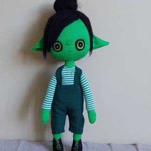Puede incluir: Muñeca verde hecha a mano con cabello negro recogido en un moño. La muñeca tiene ojos grandes y redondos y orejas puntiagudas. Viste una camisa a rayas, un peto verde y zapatos negros con cordones verdes. La muñeca está sobre un fondo neutro.