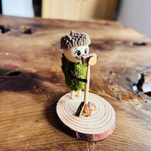 May include: A whimsical wooden figurine of a character with an acorn cap, a moss-covered body, and a wooden mallet. The figure stands on a small, round wooden base. The character has a smiling face and is holding the mallet.