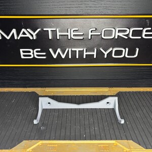 May include: A gray, 3D-printed stand with a curved design, resting on a textured surface. Above, a black sign with yellow trim displays the text "MAY THE FORCE BE WITH YOU" in white, a reference to a popular science fiction franchise.