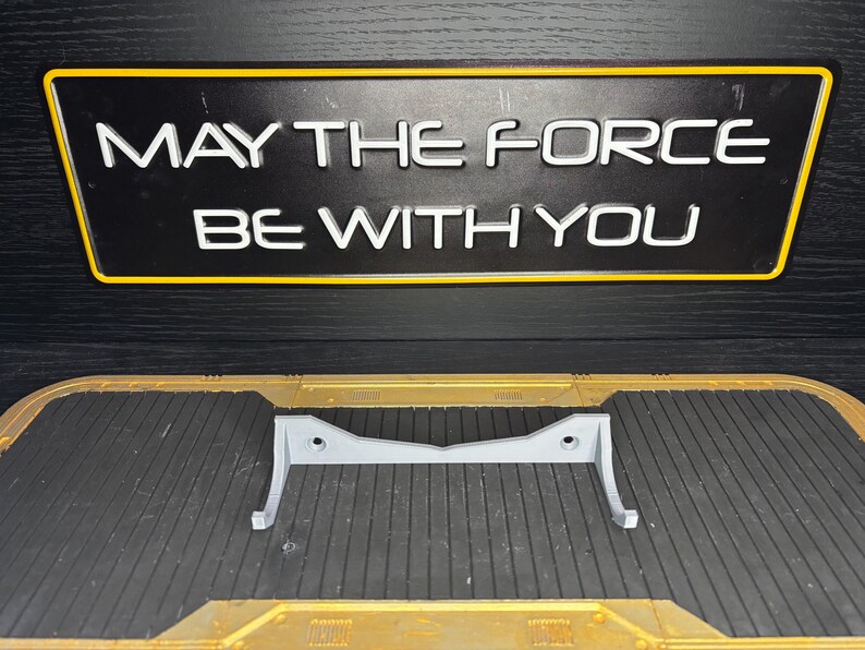 May include: A gray, 3D-printed stand with a curved design, resting on a textured surface. Above, a black sign with yellow trim displays the text "MAY THE FORCE BE WITH YOU" in white, a reference to a popular science fiction franchise.