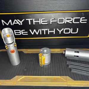 May include: A silver lightsaber replica with orange and yellow accents, disassembled, next to a sign that reads "MAY THE FORCE BE WITH YOU". The lightsaber is on a black and gold tray.
