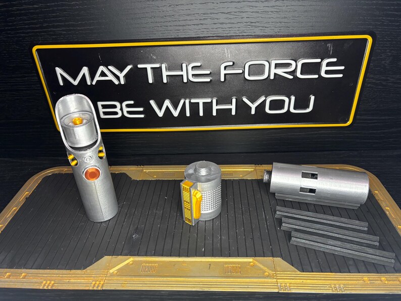 May include: A silver lightsaber replica with orange and yellow accents, disassembled, next to a sign that reads "MAY THE FORCE BE WITH YOU". The lightsaber is on a black and gold tray.