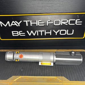 May include: A silver and black lightsaber prop with yellow accents and a red button, resting on a textured surface. Above it, a black sign with a yellow border displays the text "MAY THE FORCE BE WITH YOU" in white.