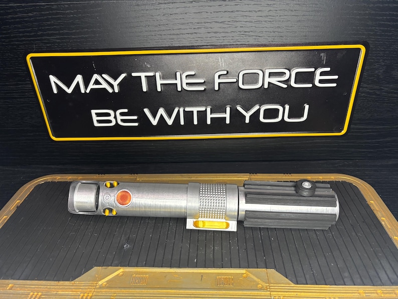 May include: A silver and black lightsaber prop with yellow accents and a red button, resting on a textured surface. Above it, a black sign with a yellow border displays the text "MAY THE FORCE BE WITH YOU" in white.