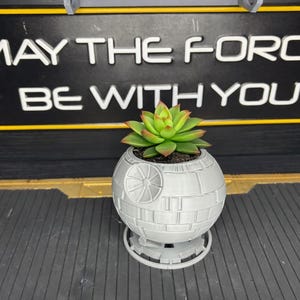 Puede incluir: Una maceta gris impresa en 3D con forma de Estrella de la Muerte con una planta suculenta. La maceta se asienta sobre una base a juego. El fondo presenta un letrero negro con el texto "MAY THE FORCE BE WITH YOU" en blanco.