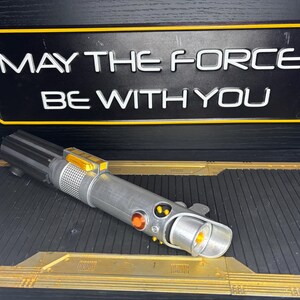 May include: A silver and black lightsaber prop with orange and yellow accents, resting on a black and gold surface. Above it, a black sign with the text "MAY THE FORCE BE WITH YOU" in white letters.