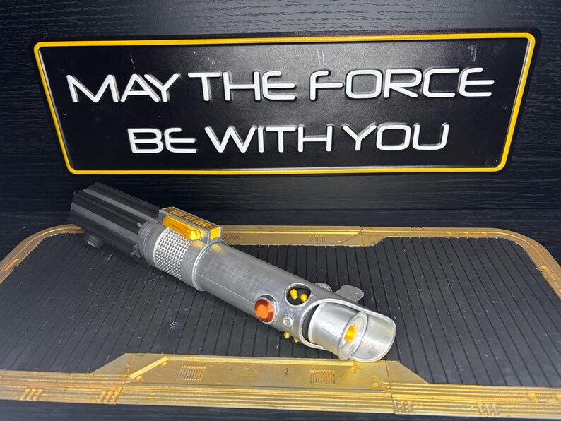 May include: A silver and black lightsaber prop with orange and yellow accents, resting on a black and gold surface. Above it, a black sign with the text "MAY THE FORCE BE WITH YOU" in white letters.