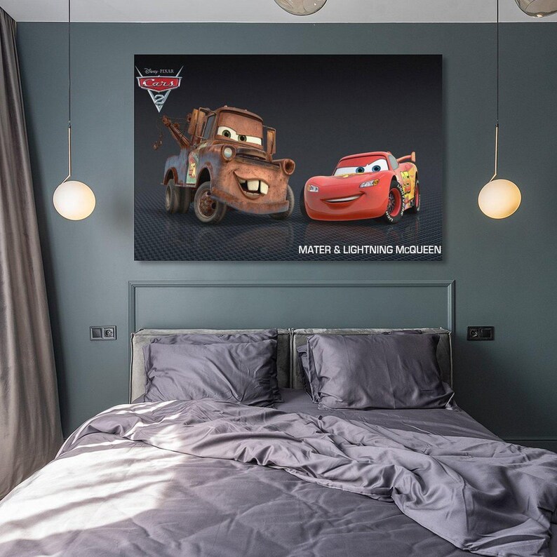 May include: A framed canvas print featuring the animated characters Mater and Lightning McQueen from the Disney Pixar Cars 2 movie. The print has a dark background with the text "MATER & LIGHTNING McQUEEN". The print is hanging above a bed with gray bedding.