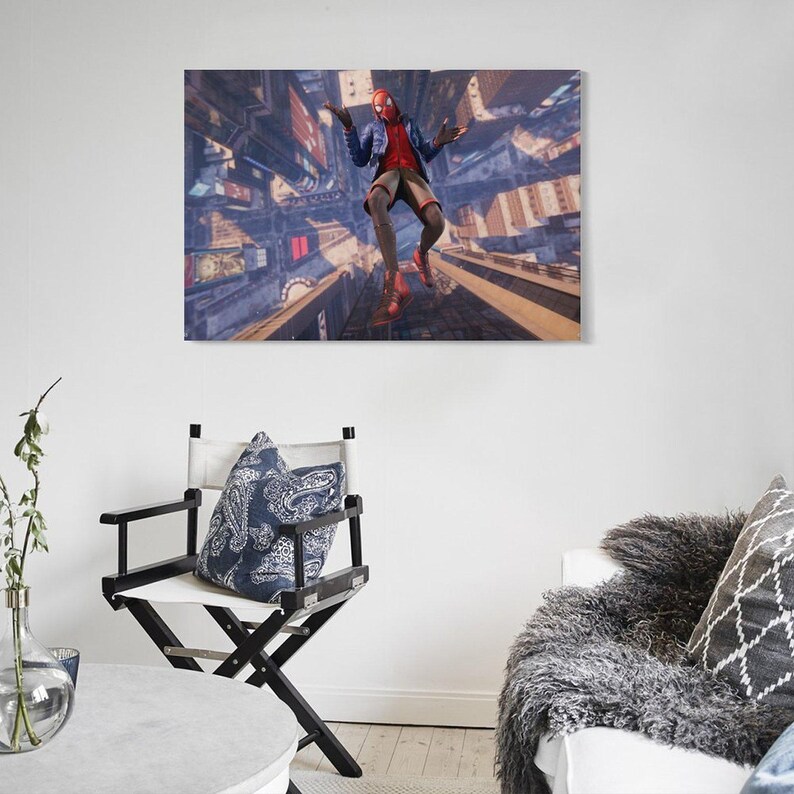 Puede incluir: Impresi&oacute;n en lienzo enmarcada que representa a un superh&eacute;roe con una m&aacute;scara roja y negra, camisa roja, chaqueta azul y pantalones cortos color canela, en el aire sobre un paisaje urbano. La obra de arte se exhibe en una pared blanca sobre una silla de director en blanco y negro.