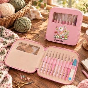 May include: A pink zippered crochet hook case featuring a floral and mushroom illustration with the name Fidelia printed below. The case is open to reveal a set of pink-handled crochet hooks and a blue-handled tool held in elastic loops.
