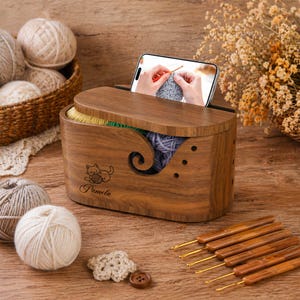 May include: A wooden yarn storage box with a lid, featuring a cat and yarn design and the name "Pamela". The box holds various coloured yarn and has a slot for a phone displaying a knitting tutorial. Crochet hooks and yarn balls are also visible.