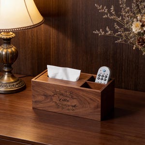 May include: A dark brown wooden tissue box with a compartment for a remote control. The box has the name "Lottie" engraved on the front. White tissues are visible in the top compartment. A lamp and flowers are in the background.