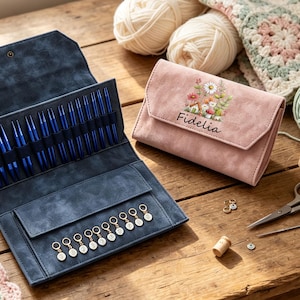 May include: A dark blue fabric case containing a set of metallic blue knitting needles and a row of small circular stitch markers. Beside it sits a closed dusty pink suede-like wallet featuring a floral illustration and the name Fidelia.
