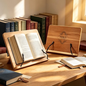 May include: Two light-toned wooden book stands with black metal page holders. One stand holds an open book, while the other is closed and features a laser-engraved diamond-shaped floral wreath design with the text M&E and the date 9.13.2025.
