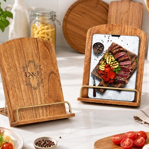 May include: Two wooden recipe holders with gold-coloured metal supports. One displays the initials "D&E" and the date "10.13.2025". The other holds a tablet showing a meal of grilled vegetables and steak. The holders are on a white surface.