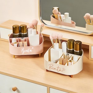May include: Two cosmetic organizers in soft pink and cream, each with multiple compartments and wooden legs. The organizers hold makeup brushes, bottles, and other beauty products. The name "Beata" is written on the front of each organizer.