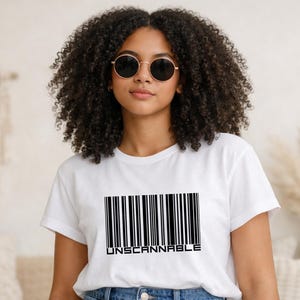 Könnte beinhalten: Ein weißes T-Shirt mit einem schwarzen Barcode-Design und dem Wort "UNSCANNABLE" darunter. Die Person auf dem Bild trägt eine Sonnenbrille und eine blaue Jeans.