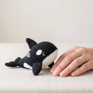 May include: A handmade crochet orca whale plushie. The toy features a black body with white patches and a white belly. The orca has a small dorsal fin and a friendly expression. The plushie is lying on a neutral surface.