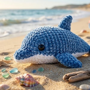 May include: A handmade blue crochet dolphin toy with a white belly, lying on a sandy beach. The scene includes seashells, sea glass, and a small yellow bucket. The ocean is visible in the background.